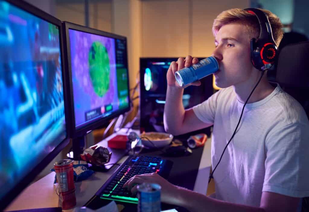 A person wearing headphones plays a video game on a dual monitor setup while sipping gamer fuel drinks, with snacks and other cans scattered across the desk.