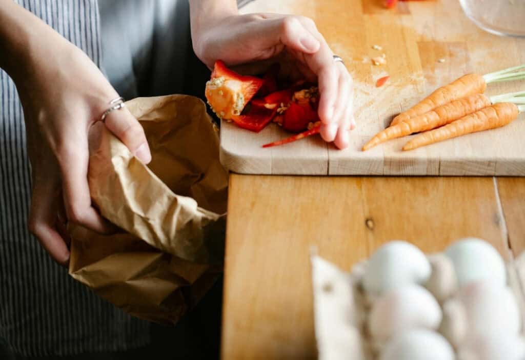 A person pushes red bell pepper scraps off a cutting board into a brown paper bag, with carrots and eggs also visible on the wooden table.
