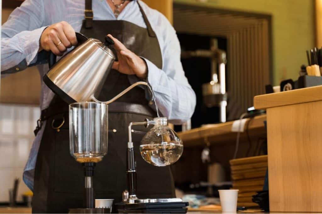 On international coffee day, a person in an apron pours hot water from a kettle into a siphon coffee maker at the café counter.