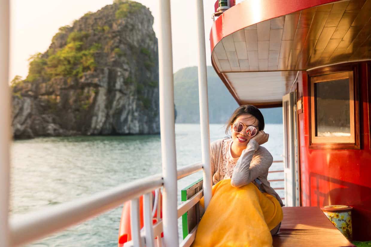 A woman wearing sunglasses and a yellow blanket sits on a boat deck, smiling, with rocky islands and water in the background.