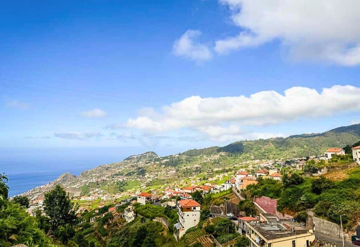 A hillside town on Madeira Island with clusters of white and red-roofed buildings overlooks the sea under a blue sky with scattered clouds.