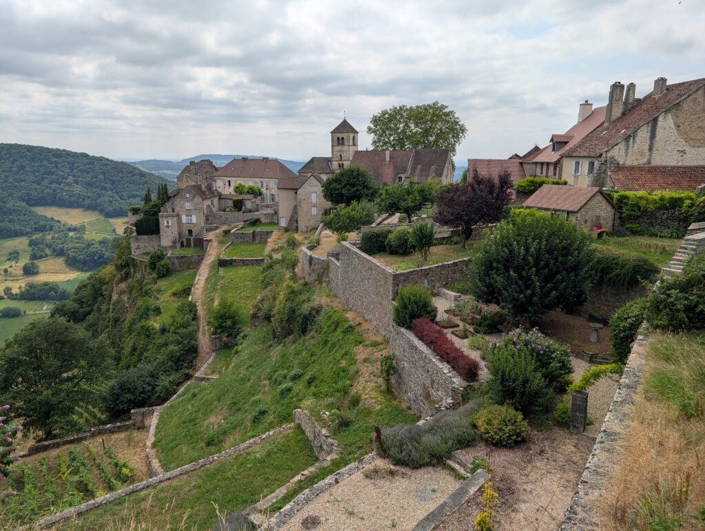 Hilltop village with stone houses, terraced gardens, and a central church tower, overlooking a green valley under a cloudy sky.