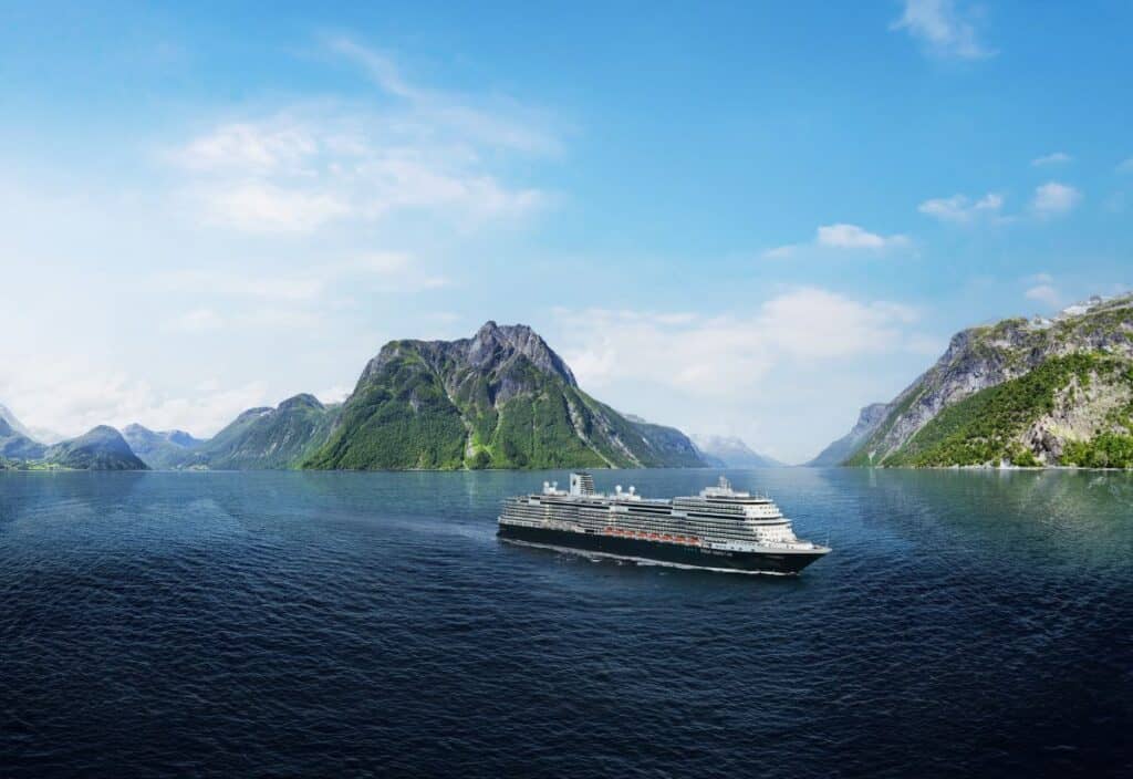 A large cruise ship sails on a calm blue sea with green mountains and a partly cloudy sky in the background.