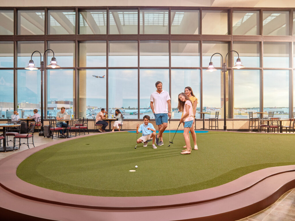 A family of four plays mini-golf indoors near large windows, with diners seated at tables in the background and an airplane visible outside.